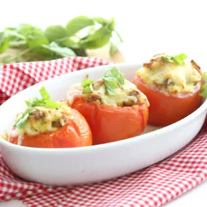 Three baked stuffed tomatoes in a white oval baking dish, filled with browned sausage and melted cheese and topped with shredded fresh basil, on a red gingham cloth
