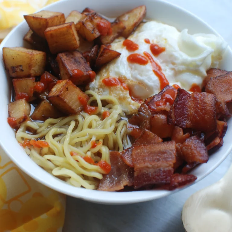 A bowl of breakfast ramen topped with crispy bacon pieces, fried potatoes, a fried egg, and a drizzle of sriracha sauce.A bowl of breakfast ramen topped with crispy bacon pieces, fried potatoes, a fried egg, and a drizzle of sriracha sauce.
