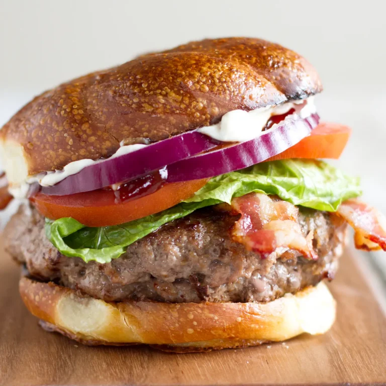 A bacon brisket burger on a toasted brioche bun stacked with a brisket-stuffed beef patty, crispy Jones Dry Aged Bacon, romaine lettuce, tomato, red onion, and garlic mayo, served on a wood cutting board.