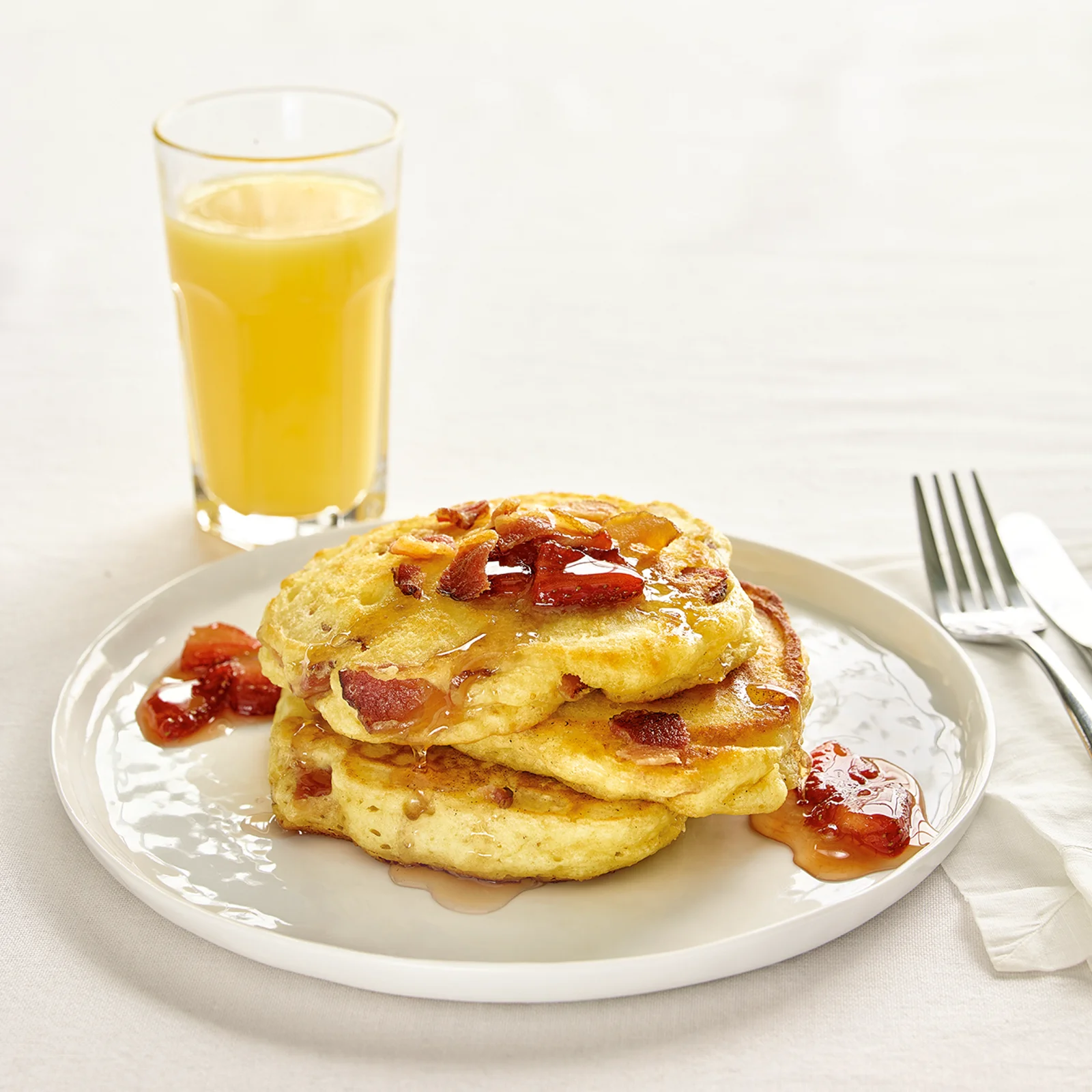 A stack of fluffy bacon pancakes with visible Jones Dry Aged Bacon crumbled throughout the batter and piled on top, drizzled with maple syrup, served on a white plate with orange juice and silverware.