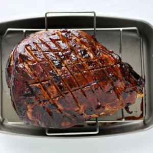 A Jones Hickory Smoked bone-in ham with a caramelized balsamic and Dijon glaze, scored in a diamond pattern, on a roasting rack in a stainless steel roasting pan.