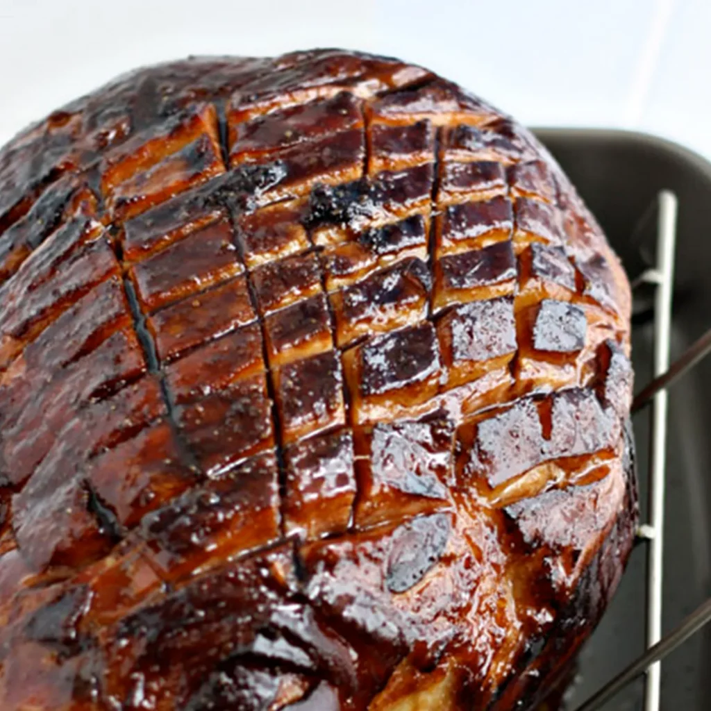 A close-up of a Jones Hickory Smoked bone-in ham with a deeply caramelized balsamic and Dijon glaze, scored in a diamond pattern, resting on a roasting rack.