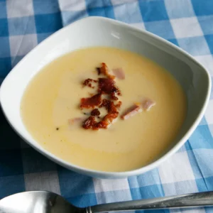 A bowl of creamy beer and cheddar soup topped with crispy Jones bacon pieces and diced Jones smoked ham, served on a blue and white checkered cloth.