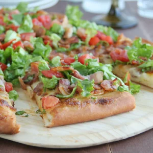 Slices of BLT pizza on a wood serving board topped with shredded lettuce, diced tomatoes, and crumbled Jones Dry Aged Bacon on a golden mayo-brushed crust.