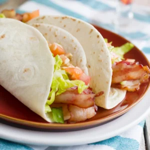 Two BLT tacos in flour tortillas filled with crispy Jones Dry Aged Bacon, shredded lettuce, and diced tomatoes, served on a brown plate with a blue striped cloth.