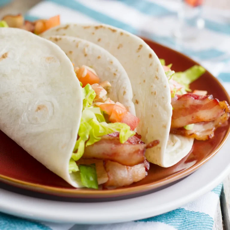 Two BLT tacos in flour tortillas filled with crispy Jones Dry Aged Bacon, shredded lettuce, and diced tomatoes, served on a brown plate with a blue striped cloth.