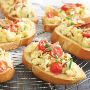 Toasted Italian bread slices topped with scrambled eggs, crispy Jones bacon, halved grape tomatoes, and fresh chives, arranged on a wire cooling rack.
