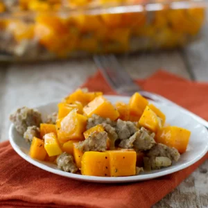 A white plate of butternut squash and sausage bake with tender cubed squash and crumbled Jones pork sausage seasoned with sage, served on an orange cloth with the full baking dish in the background.