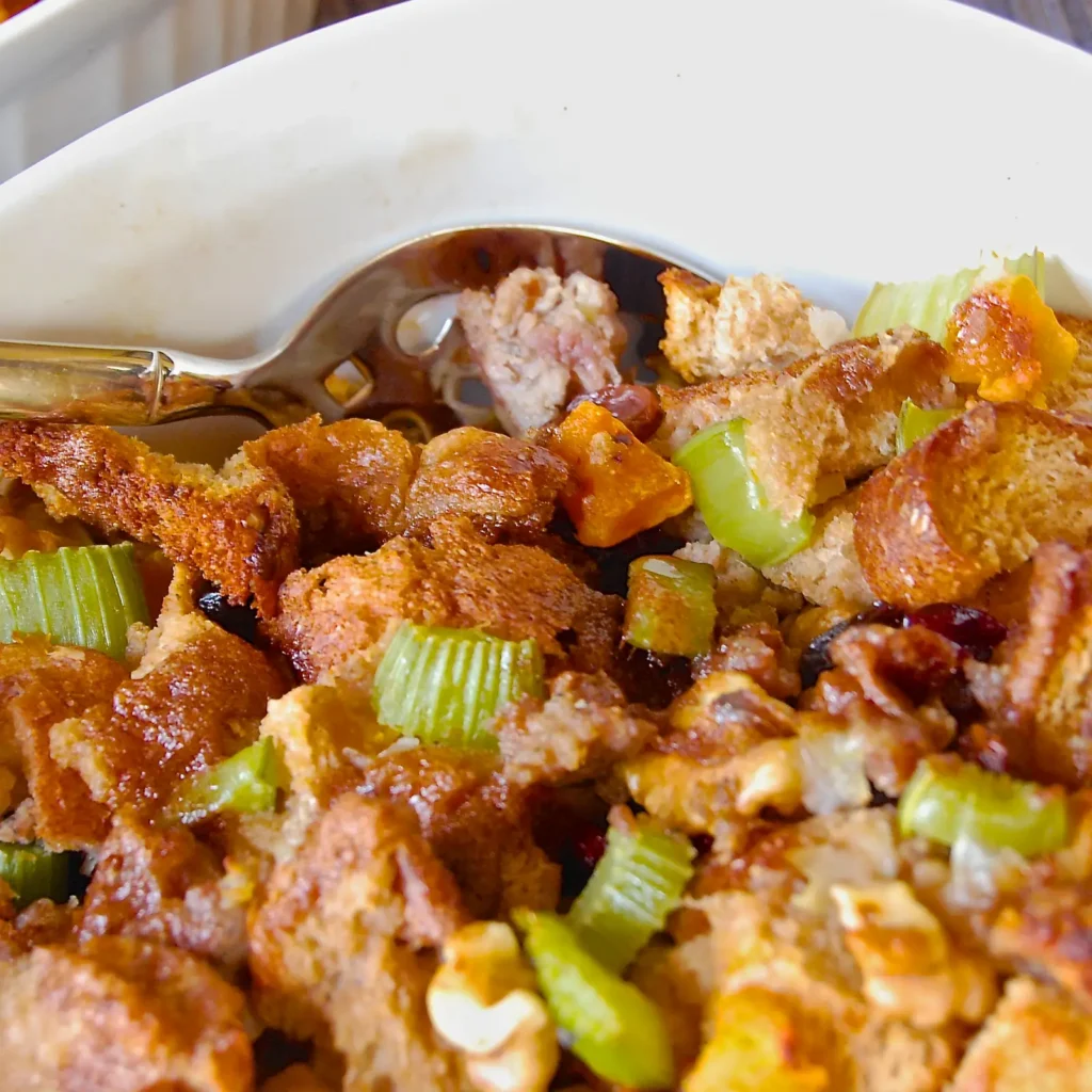 A close-up of butternut squash stuffing with Jones pork sausage, toasted bread cubes, celery, roasted squash, dried cranberries, and walnuts in a white baking …A close-up of butternut squash stuffing with Jones pork sausage, toasted bread cubes, celery, roasted squash, dried cranberries, and walnuts in a white baking dish with a serving spoon.