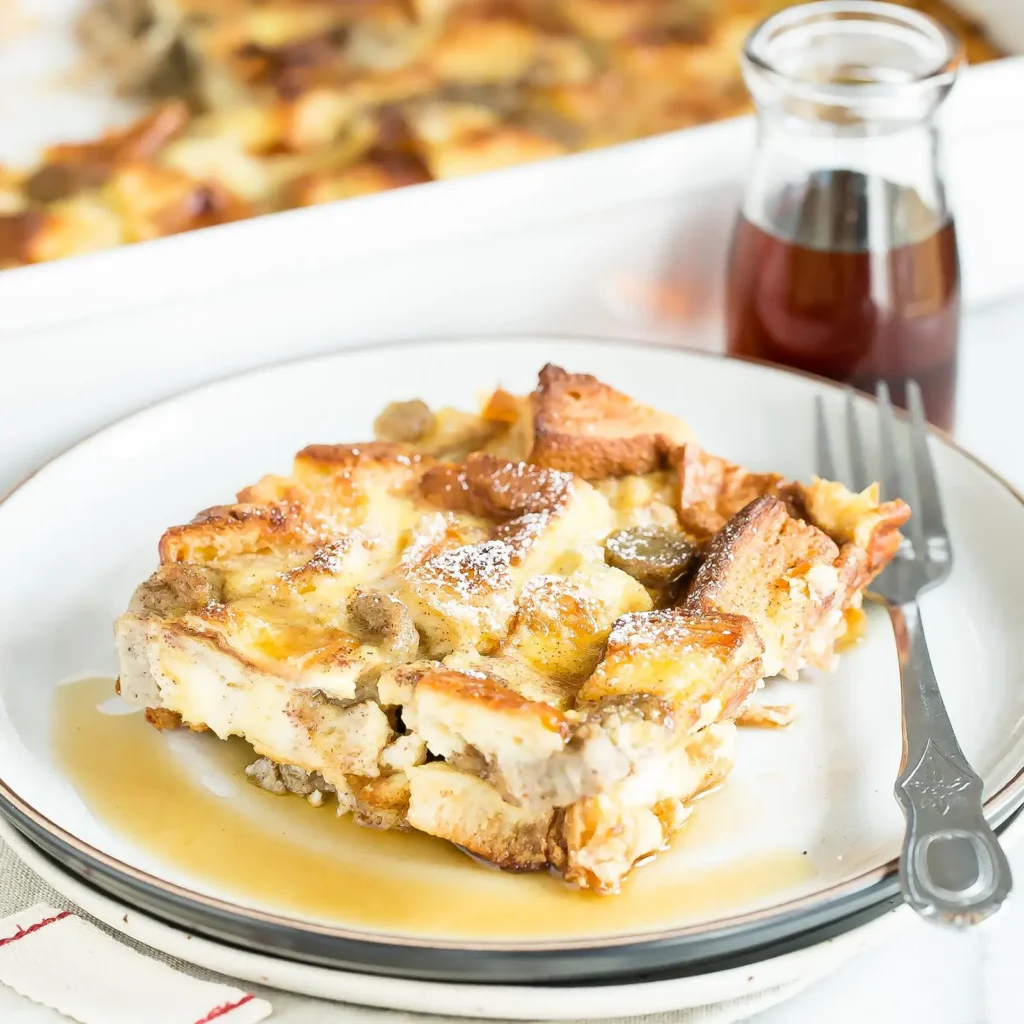 A serving of cinnamon French toast bake with Jones pork sausage baked into a golden egg custard with gluten free bread cubes, dusted with powdered sugar and drizzled with maple syrup, with the full baking dish and a maple syrup jar in the background.