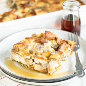 A serving of cinnamon French toast bake with Jones pork sausage baked into a golden egg custard with gluten free bread cubes, dusted with powdered sugar and drizzled with maple syrup, with the full baking dish and a maple syrup jar in the background.