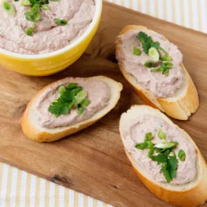 A yellow bowl of creamy braunschweiger dip made with Jones Braunschweiger, sour cream, horseradish, and green onions, served spread on sliced baguette rounds garnished with fresh parsley and green onions on a wood board.