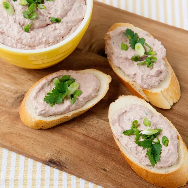 A yellow bowl of creamy braunschweiger dip made with Jones Braunschweiger, sour cream, horseradish, and green onions, served spread on sliced baguette rounds garnished with fresh parsley and green onions on a wood board.