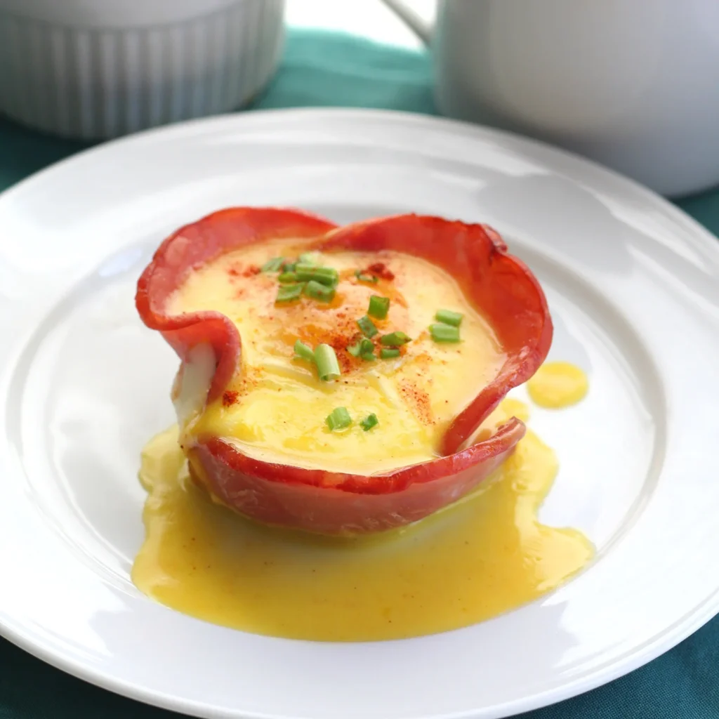 A Jones Canadian Bacon breakfast cup with a baked egg inside, drizzled with hollandaise sauce and garnished with fresh chives and paprika, served on a white plate.