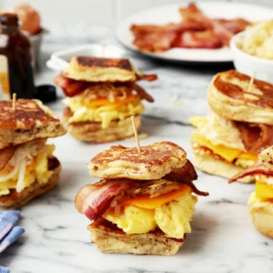 French toast breakfast sliders stacked with scrambled eggs, cheddar cheese, hash browns, and Jones Dry Aged Bacon on toothpick-skewered Hawaiian rolls, arranged on a marble surface with a plate of bacon and scrambled eggs in the background.