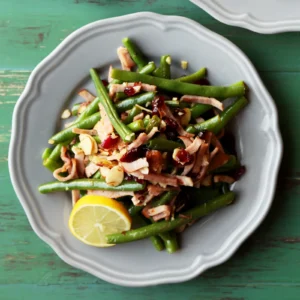 A white scalloped plate of garlic and lemon green beans tossed with julienned Jones Canadian Bacon, dried cranberries, and sliced almonds, garnished with a lemon wedge, served on a green painted wood surface.