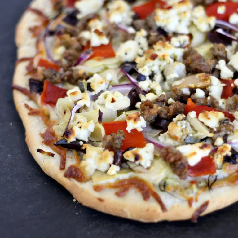 A Greek sausage pizza topped with crumbled Jones pork sausage, artichoke hearts, kalamata olives, roasted red pepper, red onion, and feta cheese on a baked crust, served on a slate surface.