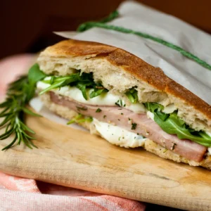 A grilled ciabatta sandwich filled with Jones Uncured Ham, fresh mozzarella, and baby arugula, served on a wood cutting board with a fresh rosemary sprig.