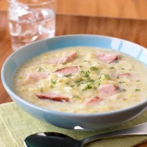 A blue bowl of ham potato leek soup with a creamy pureed leek and potato base, diced Jones Uncured Ham, and fresh dill, served on a green cloth with a spoon and water glass in the background.