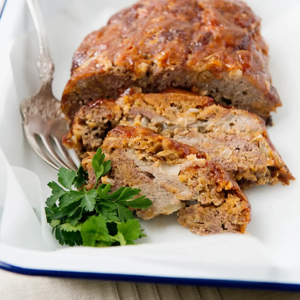 Sliced sausage meatloaf with a caramelized ketchup glaze showing a seasoned blend of Jones All Natural Sausage Roll and ground chuck, served on a white enamel platter with a fork and fresh parsley.