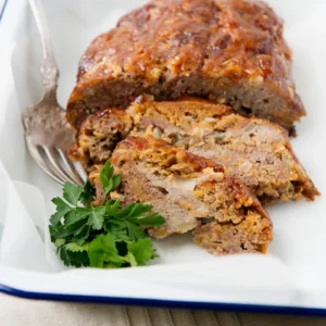 Sliced sausage meatloaf with a caramelized ketchup glaze showing a seasoned blend of Jones All Natural Sausage Roll and ground chuck, served on a white enamel platter with a fork and fresh parsley.