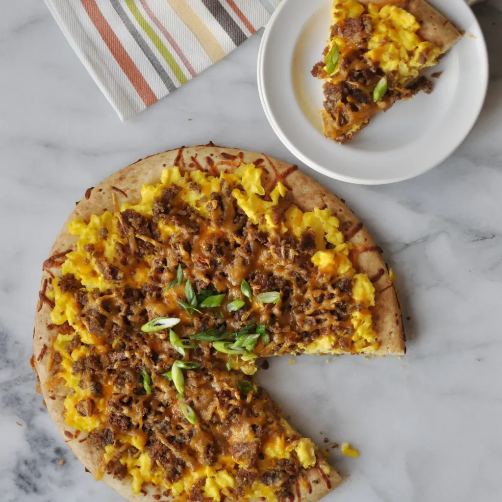 A scrapple breakfast pizza on a marble surface topped with scrambled eggs, maple-glazed crumbled Jones Scrapple, melted smoked cheddar, and sliced scallions, with a slice served on a white plate in the background.