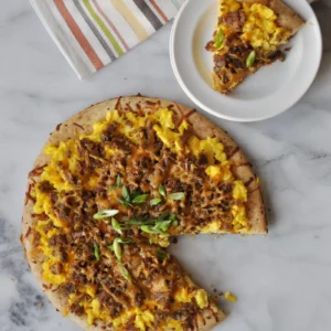A scrapple breakfast pizza on a marble surface topped with scrambled eggs, maple-glazed crumbled Jones Scrapple, melted smoked cheddar, and sliced scallions, with a slice served on a white plate in the background.
