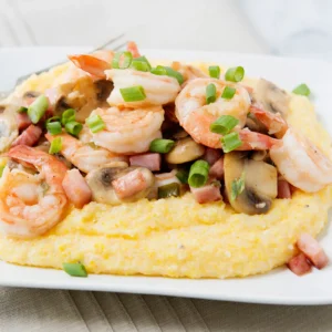 A white plate of shrimp and grits with garlic lemon shrimp, diced Jones Ham Slices, sautéed mushrooms, and green onions served over creamy stone-ground cheddar grits.