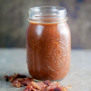 Glass mason jar filled with smoky tomato and bacon vinaigrette, garnished with crispy bacon pieces on a gray surface.