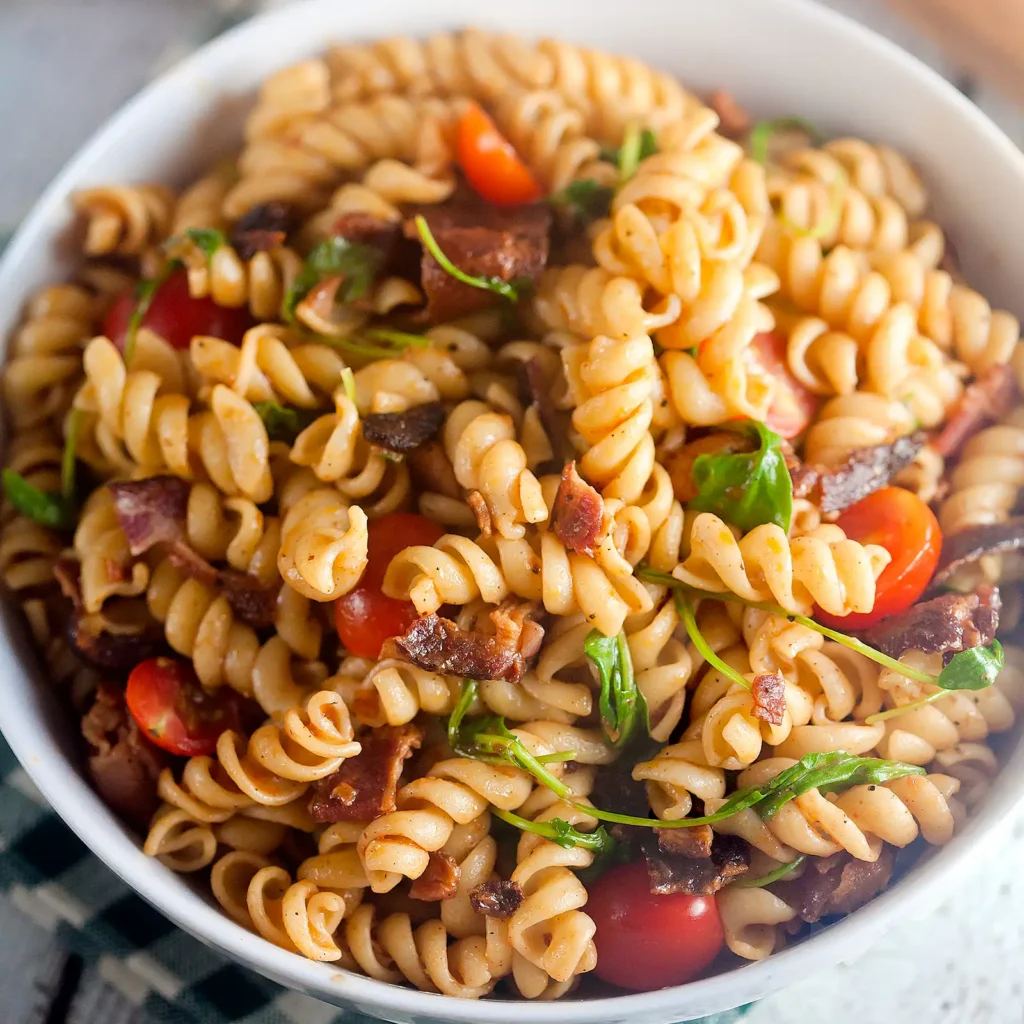 Bowl of rotini pasta salad tossed with smoked tomato and bacon vinaigrette, crispy bacon, cherry tomatoes and fresh arugula.