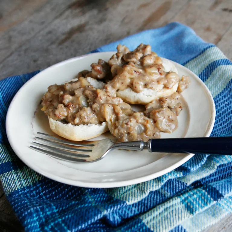 Split buttermilk biscuits topped with Southern style Jones All Natural Pork Sausage Roll and onion gravy on a white plate, served on a blue plaid cloth with a fork.
