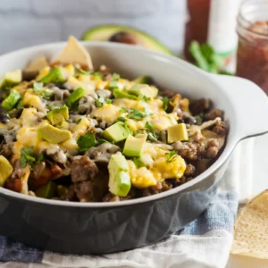 A baking dish of southwest breakfast nachos topped with melted cheese, diced avocado, and fresh cilantro, with tortilla chips, a halved avocado, and a jar of salsa in the background.