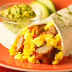 A southwest breakfast wrap filled with scrambled eggs, Jones pork sausage links, salsa, and shredded cheddar cheese in a flour tortilla, served on an orange plate with guacamole and avocado in the background.