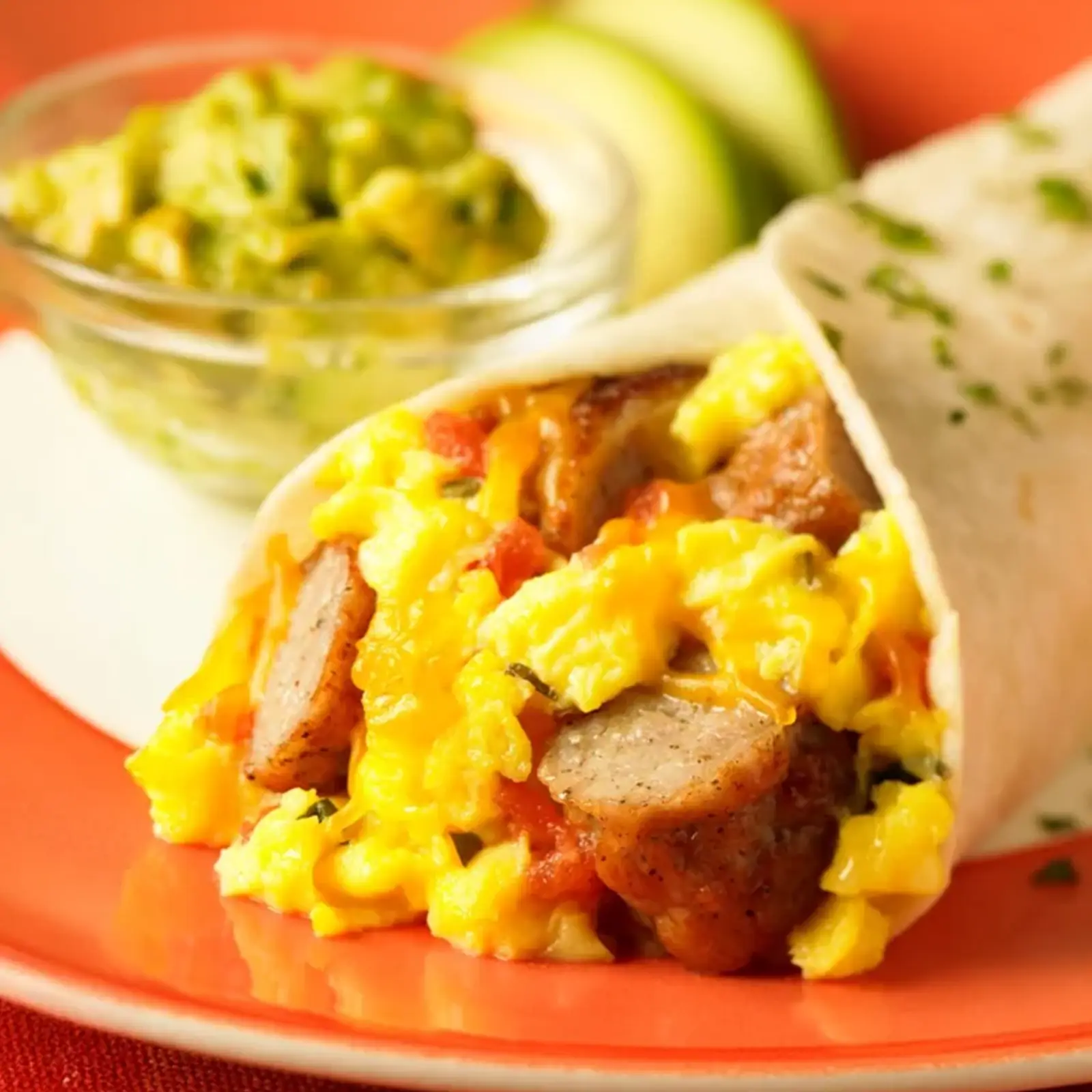 A southwest breakfast wrap filled with scrambled eggs, Jones pork sausage links, salsa, and shredded cheddar cheese in a flour tortilla, served on an orange plate with guacamole and avocado in the background.