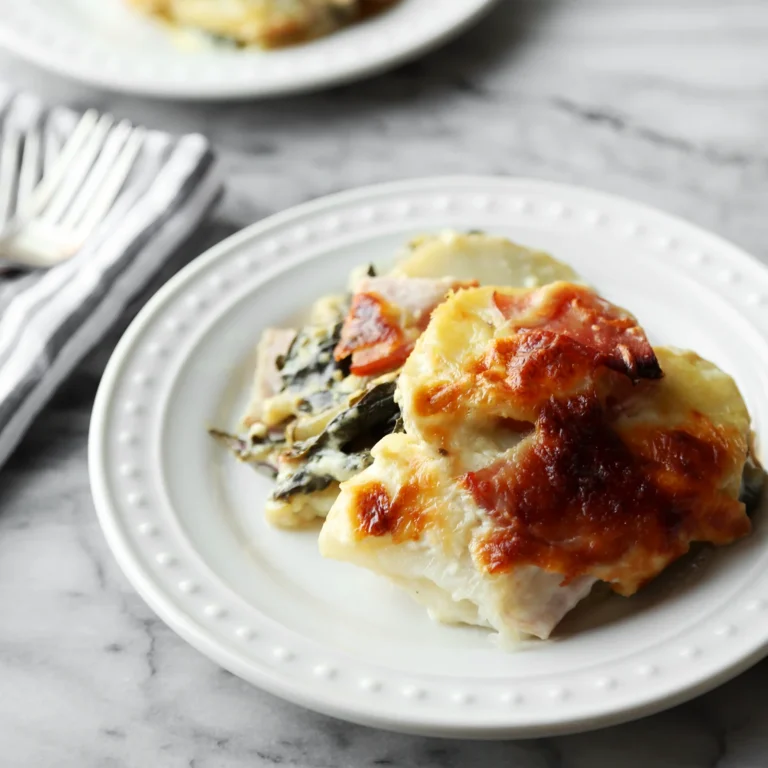 Creamy potatoes au gratin layered with Swiss chard and Canadian bacon, topped with golden brown melted cheese on a white plate.
