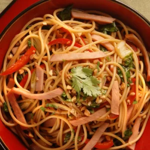 A red bowl of Szechuan-style lo mein with vermicelli noodles, strips of Jones Canadian Bacon, red bell pepper, bok choy, green onions, chopped peanuts, and fresh cilantro in a sesame soy broth.