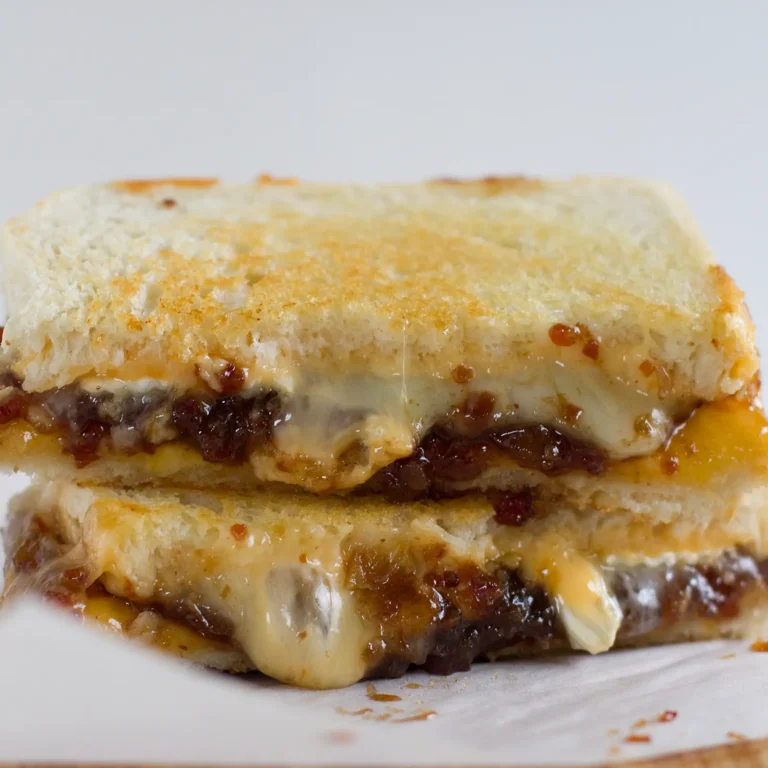 A grilled cheese sandwich cut in half and stacked, showing layers of melted brie, cheddar, and American cheese with Jones bacon jam on golden buttered bread, served on parchment paper.