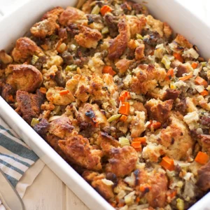 Overhead view of baked sausage stuffing in a white rectangular baking dish, with golden toasted bread cubes, crumbled sausage, diced carrots, and celery