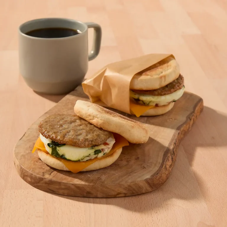 Two make-ahead breakfast sausage sandwiches on toasted English muffins with egg, spinach, and cheddar cheese, arranged on a wooden board with a mug of black coffee for meal prep, one sandwich wrapped in parchment paper for grab-and-go storage.