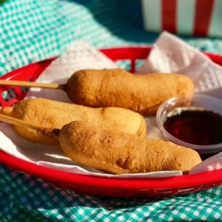 Three pancake-battered sausage links on sticks in a red basket, served with maple syrup for dipping.