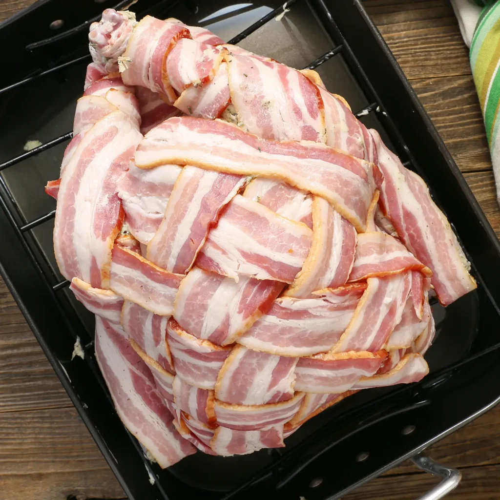 An overhead view of a whole raw turkey fully covered in a woven Jones Dairy Farm bacon lattice, resting in a black roasting pan on a wooden surface.