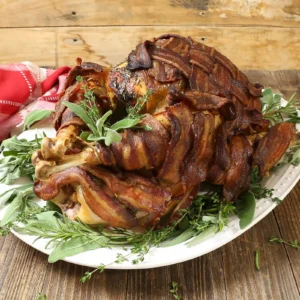 A fully roasted bacon-wrapped herb turkey with crispy, caramelized bacon on a white serving platter, garnished with fresh sage, thyme, and rosemary on a rustic wooden surface.
