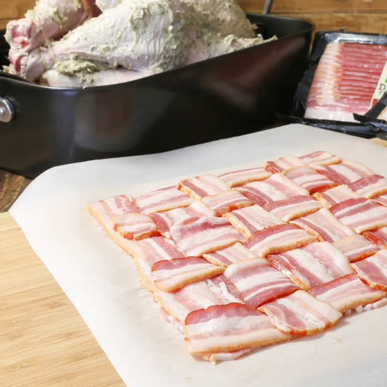 A woven bacon lattice assembled on parchment paper on a wooden cutting board, with a herb butter-coated turkey in a roasting pan and a package of Jones Dairy Farm bacon visible in the background.