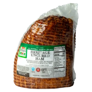 Jones Dairy Farm All Natural Heritage Uncured Half Ham in packaging. Fully cooked and hickory smoked, with a deep mahogany color and crosshatch scoring visible through the clear vacuum-sealed bag. Green and white label reads "All Natural, Hickory Smoked, Heritage Uncured Ham.