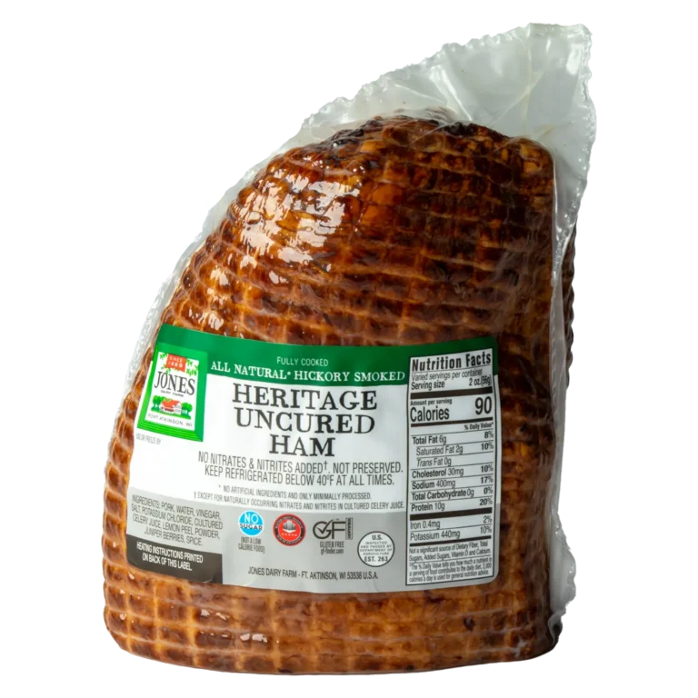 Jones Dairy Farm All Natural Heritage Uncured Half Ham in packaging. Fully cooked and hickory smoked, with a deep mahogany color and crosshatch scoring visible through the clear vacuum-sealed bag. Green and white label reads "All Natural, Hickory Smoked, Heritage Uncured Ham.