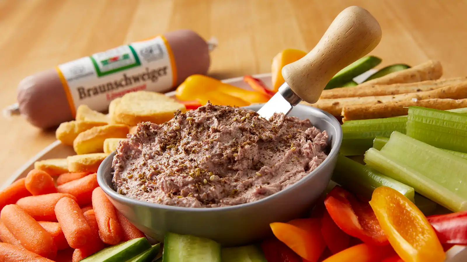 A bowl of Braunschweiger mousse dip topped with seasoning, served on a platter with carrots, celery, bell peppers, cucumber, breadsticks, and crackers, with a Jones Dairy Farm Braunschweiger Liverwurst roll in the background.