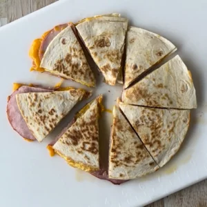 Canadian bacon quesadilla cut into six wedges and fanned out on a white cutting board, with melted cheddar cheese and thick slices of Canadian bacon visible where the filling has spilled slightly onto the board