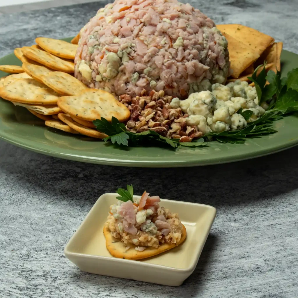 Jones Canadian bacon date cheeseball served on a cracker with blue cheese crumbles and fresh parsley, with the full cheeseball platter in the background