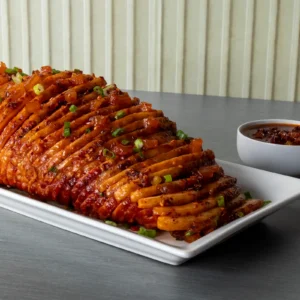 Glazed Hasselback ham with pineapple chili crisp, scallions, and a bowl of extra glaze on a white platter.