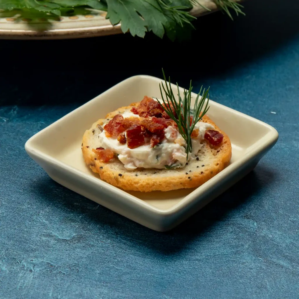 Bacon and lox cheeseball spread on a poppy seed cracker, topped with crispy Jones bacon and fresh dill